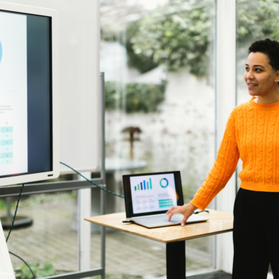 Woman in orange jumper stood presenting at a laptop