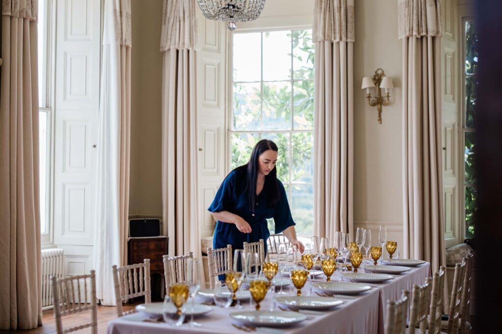 A brunette woman setting up an elegant dining table for a wedding, in a bright, sophisticated room with tall cream curtains, a chandelier, and gold-tinted glassware.