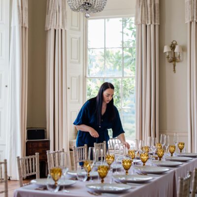 A brunette woman setting up an elegant dining table for a wedding, in a bright, sophisticated room with tall cream curtains, a chandelier, and gold-tinted glassware.